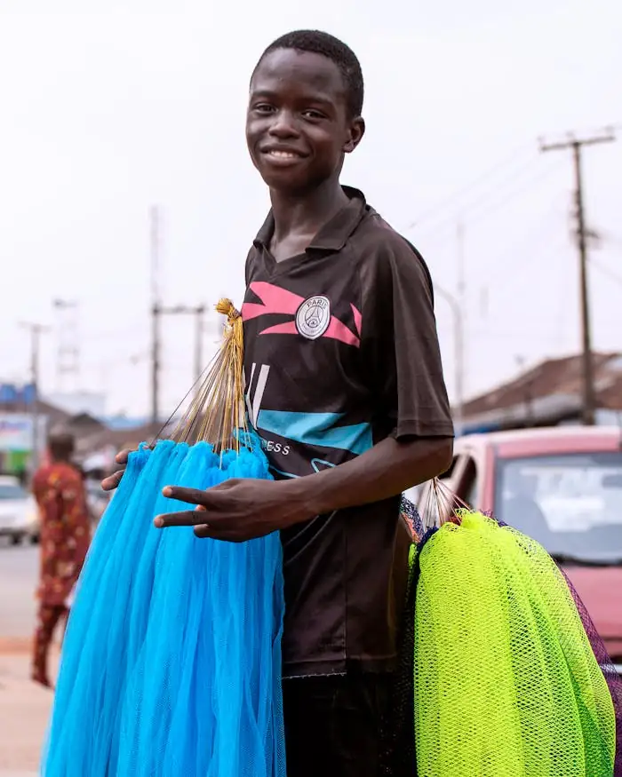 A young Nigerian vendor smiling while selling colorful nets on a busy street.