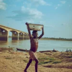 An African boy walks by a river in Nigeria holding a basin under a clear sky.