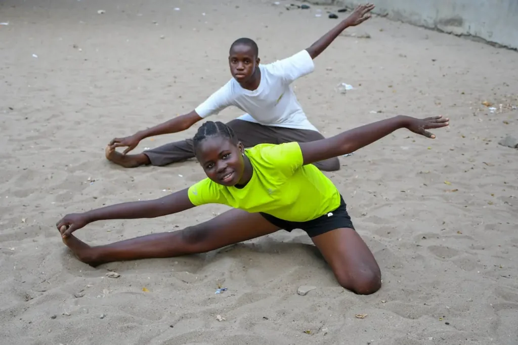 Two teenagers performing stretching exercises on sandy beach in Cotonou, Benin.