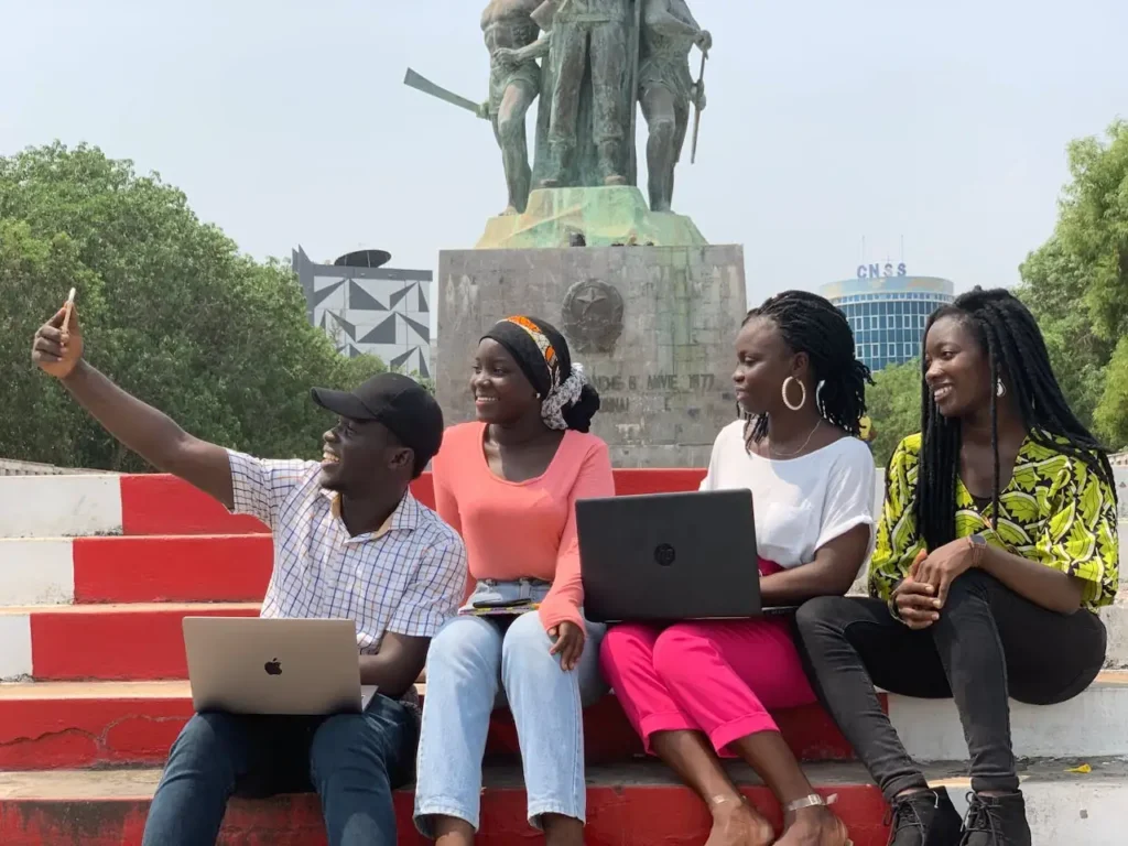 Four friends with laptops smile and take a selfie at a monument in Cotonou, Benin.